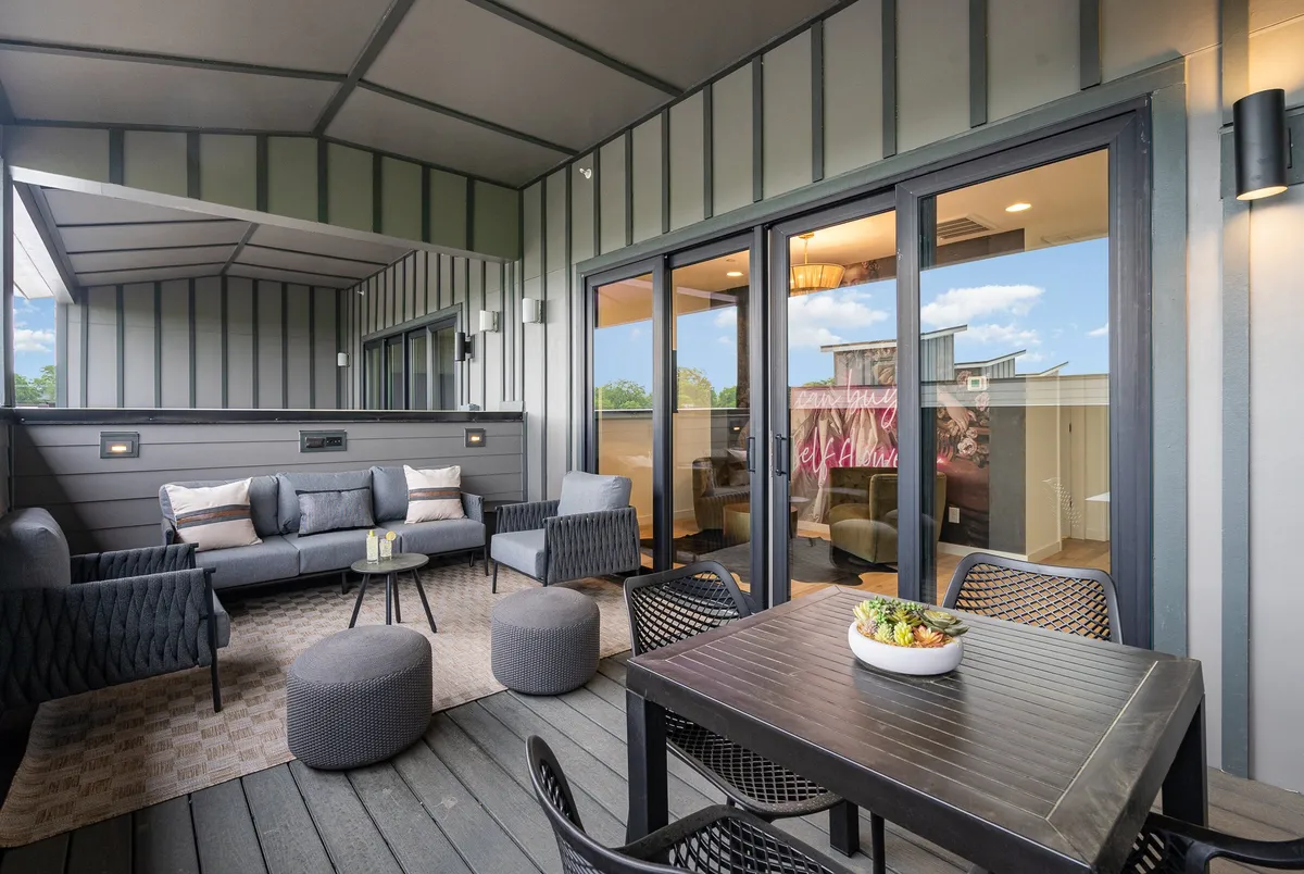 This image features an outdoor seating area with a dining table and plush seating, set just outside large glass doors. The scene is bathed in warm, natural light, suggesting a seamless connection between the indoor and outdoor living spaces. The area is inviting, comfortable, and appears ideal for group gatherings, meals, or relaxing in the fresh air.