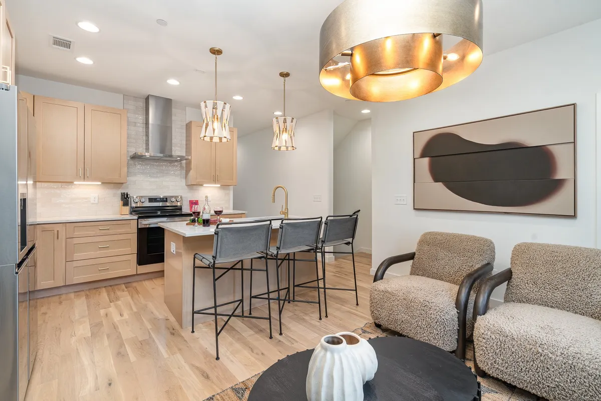 A contemporary kitchen scene with a spacious island, three bar stools, striking light fixtures, and modern artwork in the background. The island provides a central hub for cooking or conversing, while the open design visually connects the kitchen to an adjacent seating area.