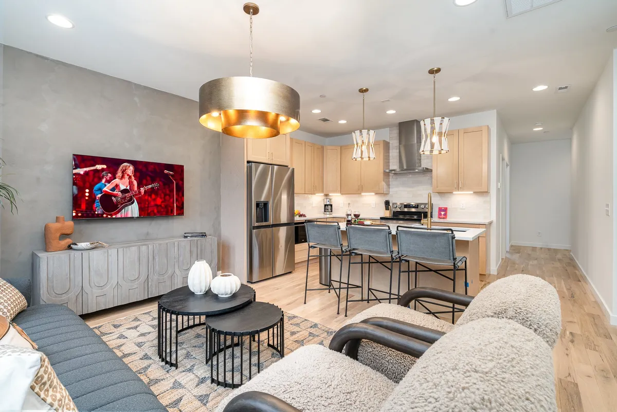 This inviting interior showcases a stylish open-concept living space with a modern kitchen island and bar stools, a cozy seating area centered around a coffee table, and a television displaying a vibrant music performance—all illuminated by elegant decorative lighting. The scene suggests a perfect blend of comfort and sophistication, ideal for relaxing or entertaining friends and family.