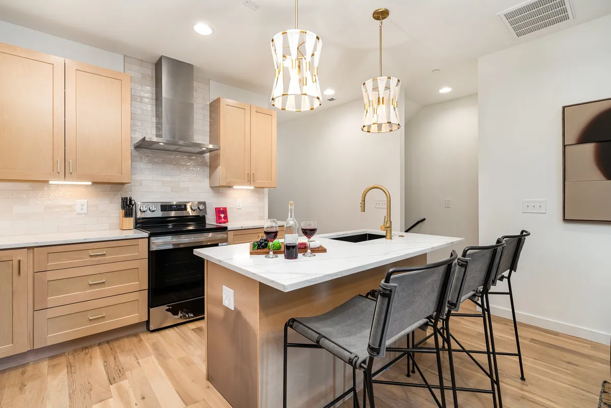 This image spotlights the kitchen island equipped with pendant lighting, bar stools, and gleaming stainless steel appliances. Wine glasses on the counter hint at social gatherings, and the overall ambience is warm and welcoming, inviting guests to cook, dine, or celebrate together.