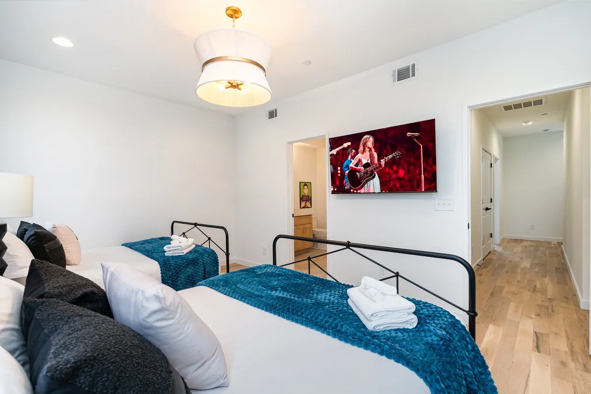 This image features two beds with textured blue blankets, a large TV mounted on the wall, and stylish chandelier lighting. The setting is modern yet cozy, suggesting a restful environment suited for relaxation, reading, or enjoying entertainment—key attributes for a guest suite or bedroom.