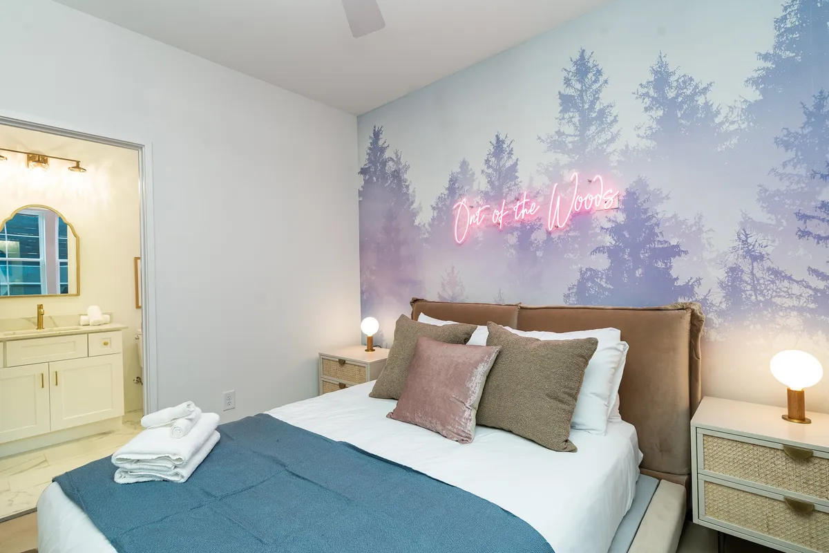 A serene bedroom highlighted by a bed with decorative pillows, a neon sign reading 'Out of the Woods,' and woodland-themed wall mural. Simple side tables with lamps flank the bed, enhancing the sense of a tranquil retreat.